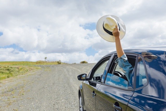 Person holding a hat out of a car window on a gravel road.
